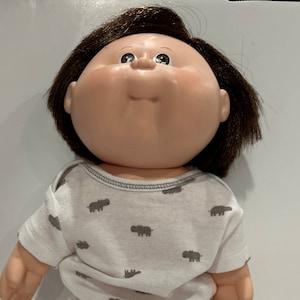 May include: A vintage doll with a round face, brown hair, and a white onesie and shorts set. The clothing is patterned with gray hippopotamus figures. The doll has a classic, nostalgic appearance.