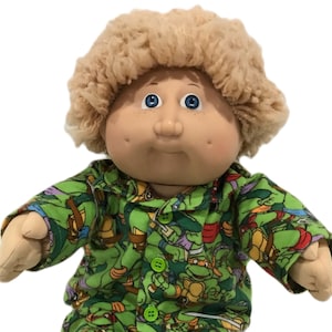 May include: A Cabbage Patch Kids doll wearing green pajamas patterned with Teenage Mutant Ninja Turtles. The doll has blonde, curly hair, blue eyes, and rosy cheeks. The pajamas have green buttons and green feet.