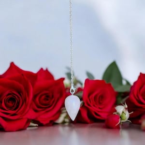May include: A white teardrop-shaped crystal pendulum on a silver chain, hanging above a cluster of red roses.