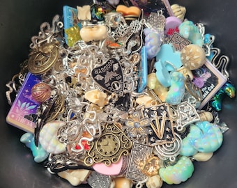 Könnte beinhalten: Eine schwarze Schüssel gefüllt mit einer Vielzahl von Charms, Perlen und Schmuckstücken. Die Charms beinhalten eine Uhr, ein Herz mit einer Fledermaus, einen Halbmond und eine Tarotkarte mit dem Text "The Moon". Die Perlen sind in Blautönen, Rosa und Weiß.