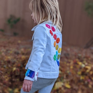 Image shows a small child wearing a white jean jacket. There are applique hexagons cascading down the back of the jacket in a rainbow hombre pattern. The jacket cuffs feature matching colored bands.