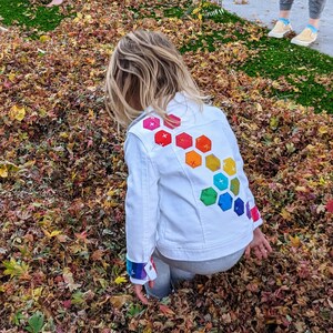 Image shows a small child wearing a white jean jacket playing in a pile of leaves. There are applique hexagons cascading down the back of the jacket and matching cuffs in rainbow colors (pink, red, orange, yellow, green, blue, purple).