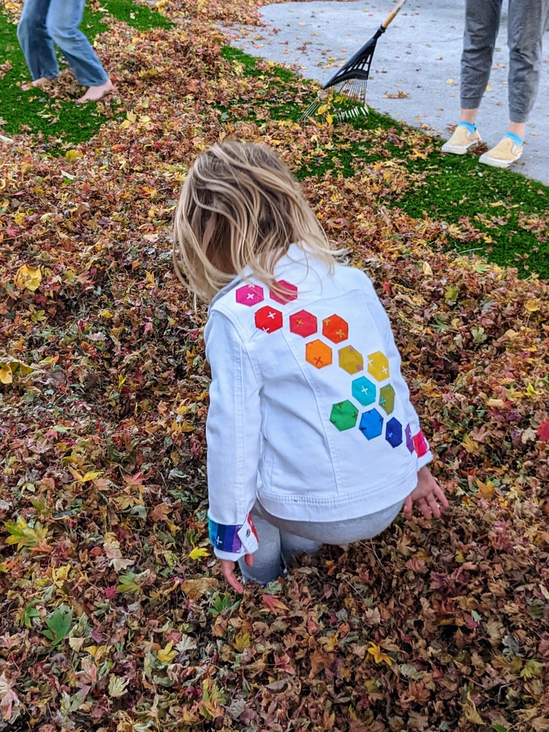 Image shows a small child wearing a white jean jacket playing in a pile of leaves. There are applique hexagons cascading down the back of the jacket and matching cuffs in rainbow colors (pink, red, orange, yellow, green, blue, purple).