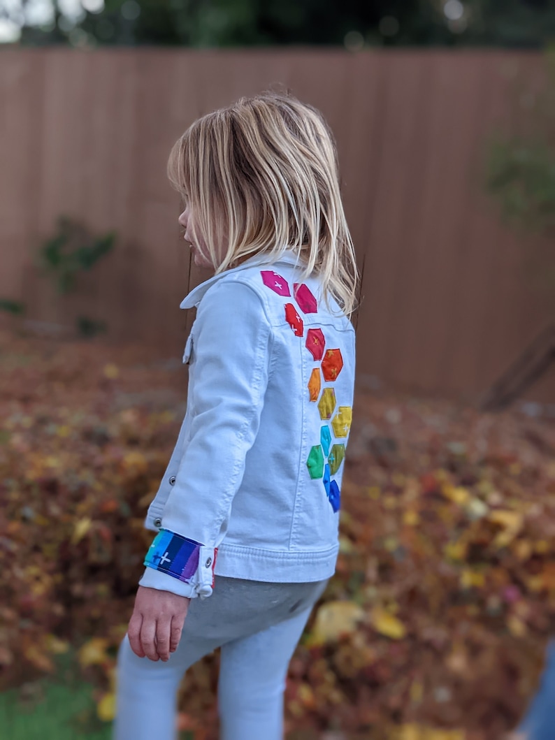 Image shows a small child wearing a white jean jacket. There are applique hexagons cascading down the back of the jacket in a rainbow hombre pattern. The jacket cuffs feature matching colored bands.