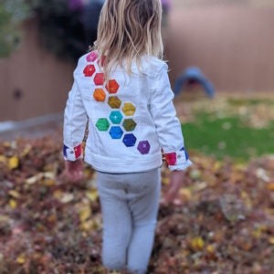 Image shows a small child standing in a pile of leaves wearing a white jean jacket. There are applique hexagons cascading down the back of the jacket in a rainbow hombre pattern. The jacket cuffs feature matching colored bands.