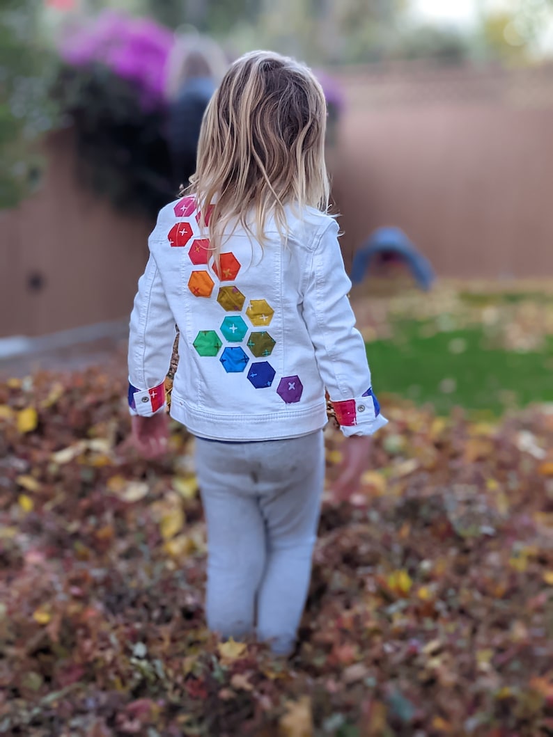 Image shows a small child standing in a pile of leaves wearing a white jean jacket. There are applique hexagons cascading down the back of the jacket in a rainbow hombre pattern. The jacket cuffs feature matching colored bands.