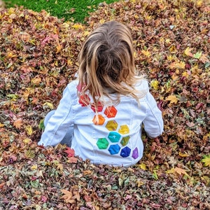 Image shows a small child wearing a white jean jacket sitting in a pile of leaves. There are applique hexagons cascading down the back of the jacket in a rainbow colors (pink, red, orange, yellow, green, blue, purple).