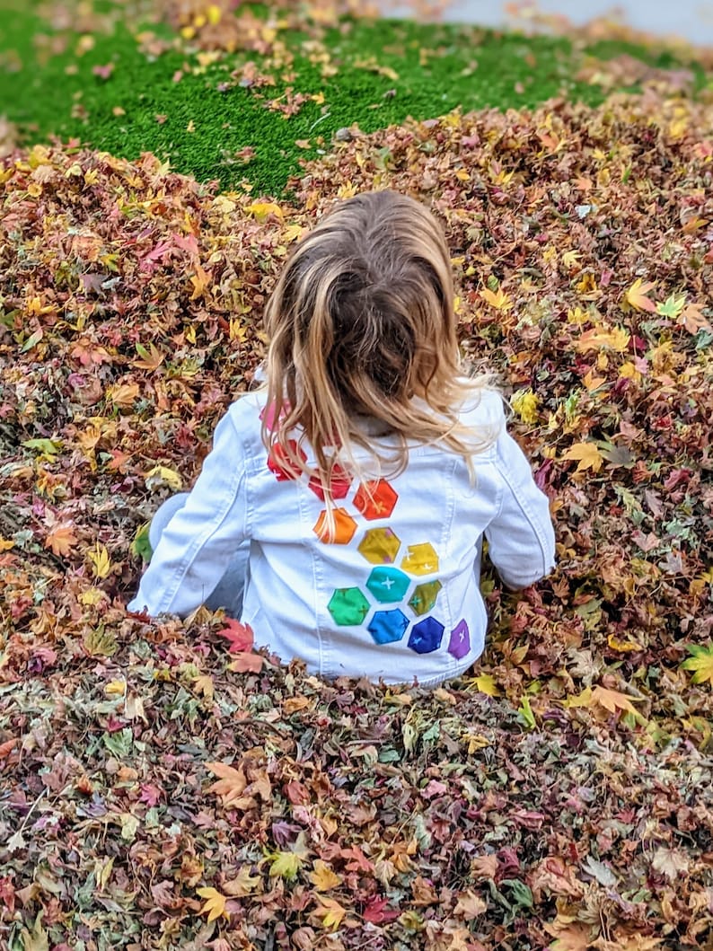 Image shows a small child wearing a white jean jacket sitting in a pile of leaves. There are applique hexagons cascading down the back of the jacket in a rainbow colors (pink, red, orange, yellow, green, blue, purple).