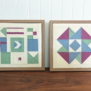 May include: Two framed pieces of quilted fabric art. The left piece features a geometric pattern in shades of green, blue, and pink. The right piece features a starburst pattern in shades of green, blue, and pink.