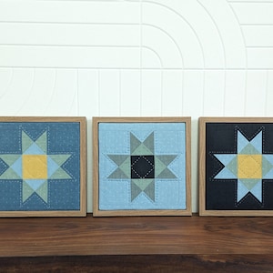 May include: Three framed fabric wall art pieces with a starburst design. The starburst is made of blue, gray, and yellow fabric. The frames are a light brown wood.