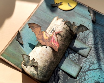 May include: A Halloween card featuring a pop-up tower with bats flying around a full moon. The tower is made of stone and has a witch and a man on top. The bats are black and have a spooky look.