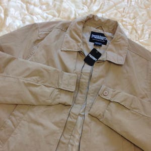 Puede incluir: Una chaqueta beige con cremallera, cuello y dos bolsillos. La chaqueta tiene una etiqueta de marca que dice "Steele & Brandt".