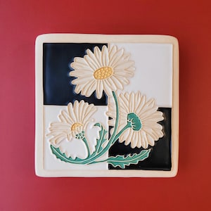 May include: Square ceramic tile featuring a daisy floral design. The tile has a checkerboard pattern of black and white squares. The daisies are cream-colored with yellow centers and green stems and leaves. The tile has a cream-colored border.