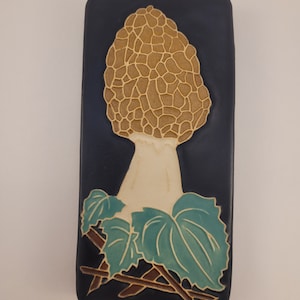May include: A rectangular, dark blue decorative tile featuring a detailed, tan morel mushroom design. Below the mushroom are light green leaves and brown stems. The mushroom has a textured, honeycomb-like pattern.