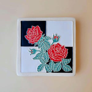 May include: Square ceramic tile featuring a red rose design with green leaves and buds. The background is a checkerboard pattern of black and white, framed by a white border. A decorative piece for home decor.