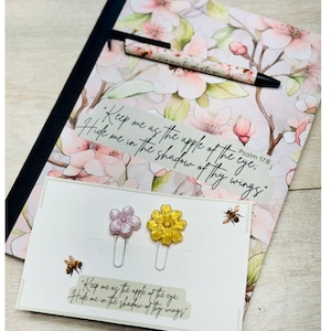 May include: Two floral-themed paper clips, one pink and one yellow, with a white card that reads "Keep me as the apple of the eye. Hide me in the shadow of thy wings."  Psalm 17:8