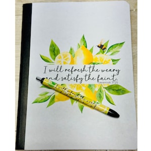 May include: A white notebook with a black spine and a pen with a lemon design. The notebook has a watercolor illustration of lemons and a bee. The text on the notebook reads "I will refresh the weary and satisfy the faint. Jeremiah 31:25".