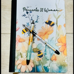 May include: A white journal with a floral design and the text "Proverbs 31 Woman" on the cover. Two pens with floral designs are on top of the journal.