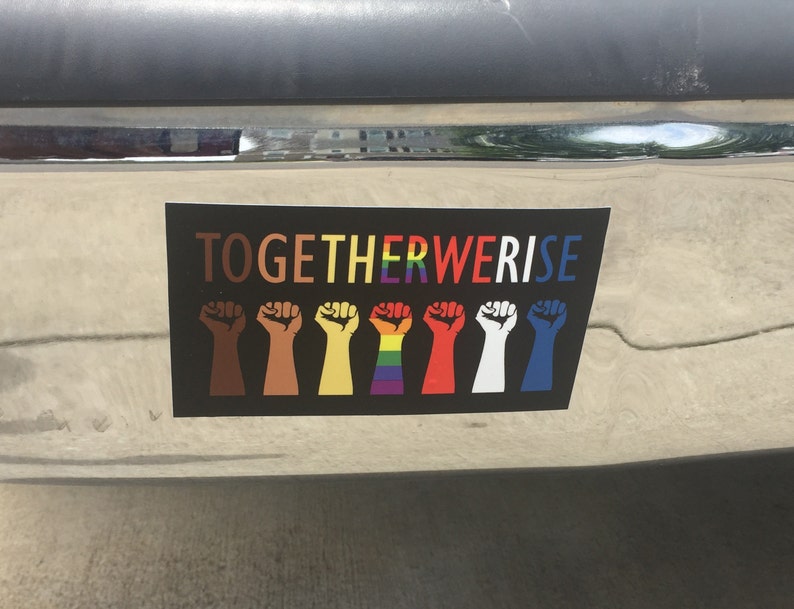 May include: A black sticker with the words "TOGETHER WE RISE" in rainbow colors. Below the text are seven raised fists in various skin tones and rainbow colors, symbolizing unity and diversity.