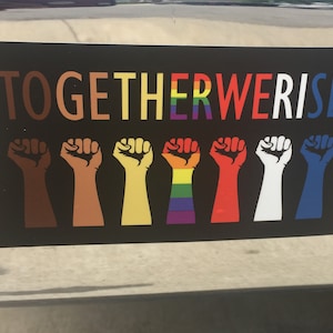May include: Black sticker with the text "TOGETHER WE RISE" in various colors. Below the text are eight raised fists in different skin tones and colors, including a rainbow-colored fist.