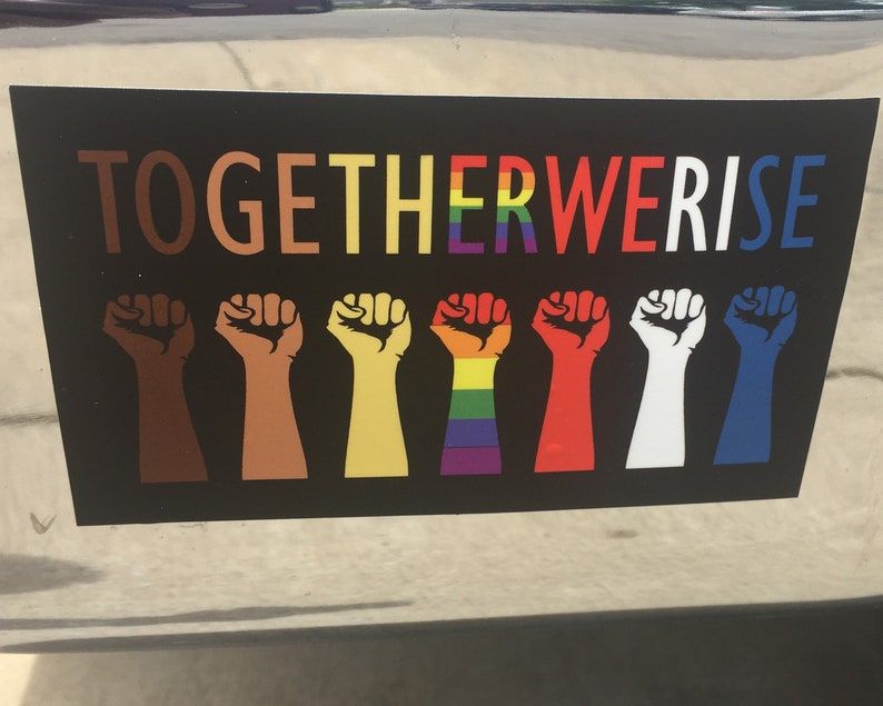 May include: Black sticker with the text "TOGETHER WE RISE" in various colors. Below the text are eight raised fists in different skin tones and colors, including a rainbow-colored fist.
