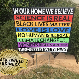 May include: A colorful yard sign with the text "In our home we believe" and a list of statements including "Science is real", "Black lives matter", "Love is love", "No human is illegal", "Climate change is real", "Women's rights are human rights", and "Kindness is everything".