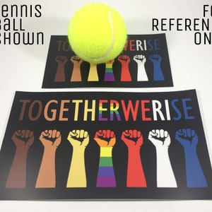 May include: Two black posters with the text "TOGETHER WE RISE" and illustrations of fists in various colors, including brown, yellow, red, white, and blue. A tennis ball is placed on top of one of the posters. The text "TENNIS BALL SHOWN" and "FOR REFERENCE ONLY" are also visible.