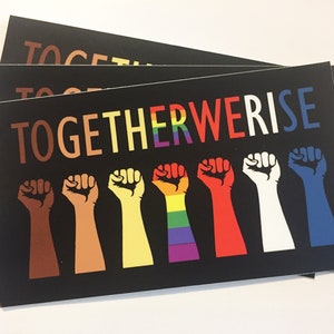 May include: Black rectangular cards with the words "TOGETHER WE RISE" in colorful letters. Below the text are seven raised fists in various skin tones and colors, including a rainbow-colored fist. The cards are stacked.