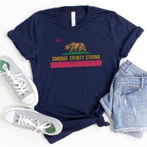 May include: A navy blue t-shirt with a distressed California flag design featuring a red star, a brown bear, and the text "Sonoma County Strong".