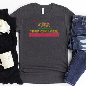 May include: A dark heather grey t-shirt with a distressed graphic of the California state flag featuring a bear and the text "Sonoma County Strong".