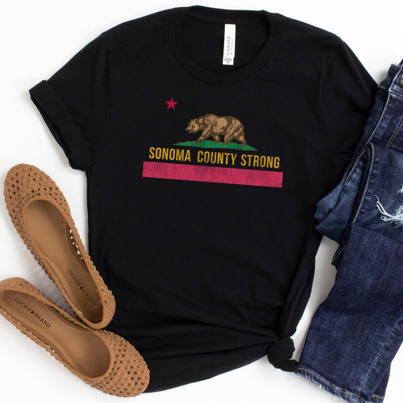 May include: Black t-shirt with a graphic of the California state flag featuring a bear and the text "Sonoma County Strong". The t-shirt is laid flat on a white surface with a pair of brown woven flats and blue jeans.