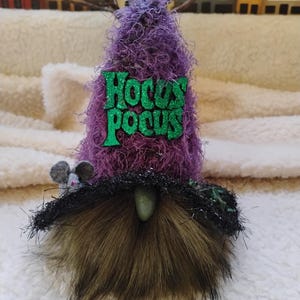 May include: A Halloween gnome with a purple, textured hat and a brown fur beard. The hat features the text "Hocus Pocus" in green glitter. A small gray mouse is perched on the gnome's head. Decorative item.