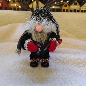 May include: A Christmas gnome figurine with a black and silver hat and coat, red mittens, and black boots with gold and red accents. The gnome has a long white beard and a pink nose, holding a book and a small bell. The background features a snowy landscape and a village.