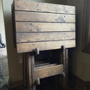 May include: A set of four dark brown wooden folding snack tables stacked vertically in a storage rack. Each table features a rectangular top made of horizontal planks with visible wood grain and small metal screws securing the surface boards.