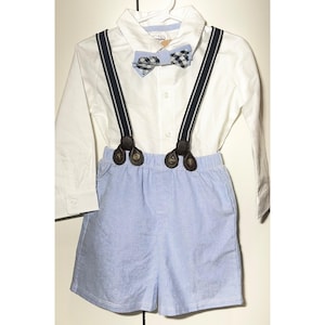 May include: A white long-sleeve shirt with a blue and white checkered bow tie and blue suspenders. The outfit includes light blue shorts. The suspenders have brown leather-like attachments.