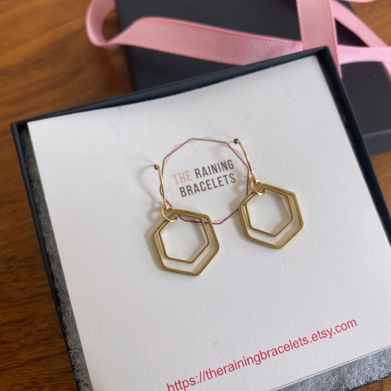 May include: Gold double hexagon dangle earrings in a white box with the text "THE RAINING BRACELETS" and the website address "https://therainingbracelets.etsy.com".