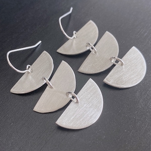 May include: A pair of silver-toned drop earrings, each featuring three vertically linked semi-circular metal pieces with a brushed finish. The earrings are suspended from curved silver-toned ear wires, creating a tiered, geometric design.