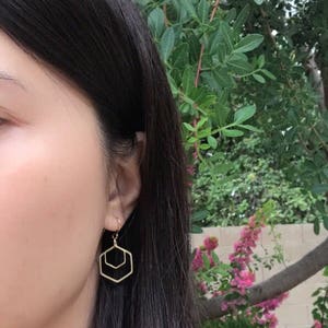 May include: Gold geometric hexagon earrings with a double-layered design. The earrings are dangling from the earlobe.