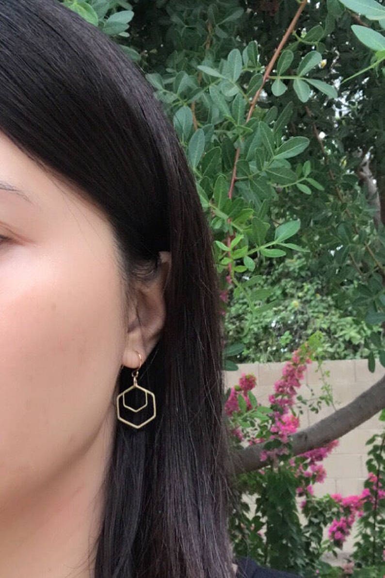 May include: Gold geometric hexagon earrings with a double-layered design. The earrings are dangling from the earlobe.