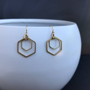 May include: A pair of gold-toned earrings with a double hexagon design. The earrings are hanging from a white surface.