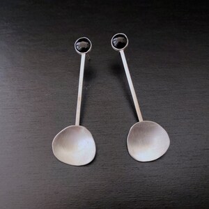 May include: A pair of silver earrings with black gemstone accents. The earrings feature a long, thin stem and a rounded, slightly oval-shaped bottom.
