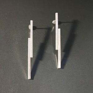May include: A pair of silver earrings with a geometric design. Each earring is made of three thin, rectangular bars stacked vertically.