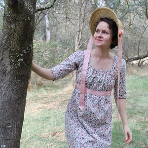 Könnte beinhalten: Ein geblümtes Kleid mit rundem Ausschnitt und Dreiviertelärmeln. Das Kleid hat einen rosa Schleifengürtel und wird mit einem Strohhut mit rosa Blume und Bändern kombiniert. Die Frau steht neben einem Baum in einer natürlichen Umgebung.