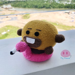 May include: A crocheted brown bear plush toy with a white smile and black eyes, holding a pink crocheted flamingo pool float. The bear is sitting on a white surface. The text "Pig Moonie" is visible in the lower right corner of the image.
