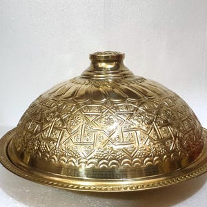 Könnte beinhalten: Eine goldfarbene Servierhaube aus Metall mit einem komplizierten, geprägten Muster. Die Haube hat eine runde Basis und einen gewölbten Deckel.