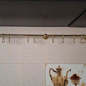 May include: A horizontal gold-toned metal rail mounted to a wall, featuring a series of matching gold-toned S-shaped hooks suspended along its length. The rail is secured by circular wall brackets at both ends and a central support point.