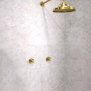 Könnte beinhalten: Ein goldfarbener Duschkopf mit rundem Sprühkopf und passendem Wandarm, ergänzt durch zwei separate goldfarbene Kreuzgriff-Ventile, die alle an einer Wand mit weißen Marmorfliesen und grauen Aderungen montiert sind.