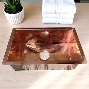 Op de afbeelding: Een koperkleurige, rechthoekige wastafel met een afvoergat. De wastafel staat op een lichtgekleurd houten oppervlak, met een kleine potplant en een stapel witte handdoeken op de achtergrond. De tekst "Kiryakoshop" is zichtbaar op de wastafel.