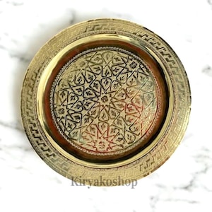May include: A round, gold-coloured metal tray with an intricate, embossed design. The tray has a raised rim with a geometric pattern.