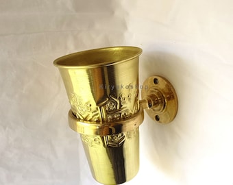 May include: A gold-colored metal toothbrush holder with an ornate design. The holder is mounted to a wall with a round base.